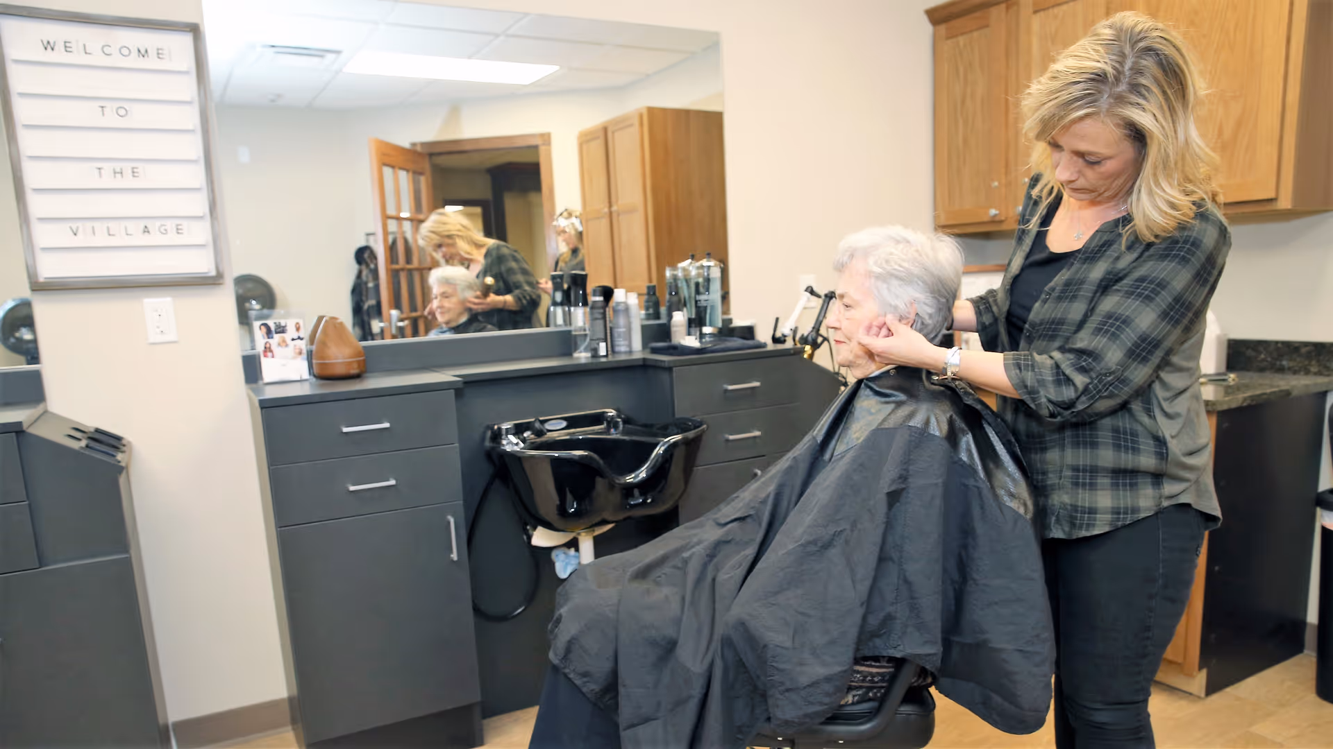 A hairstylist is cutting the hair of an elderly woman seated in a salon chair inside a hair salon area. The elderly woman is covered with a black cape. There is a large mirror on the wall reflecting the scene, and a sign on the wall reads 'WELCOME TO THE VILLAGE'. The salon has dark cabinetry and a black hair washing sink.