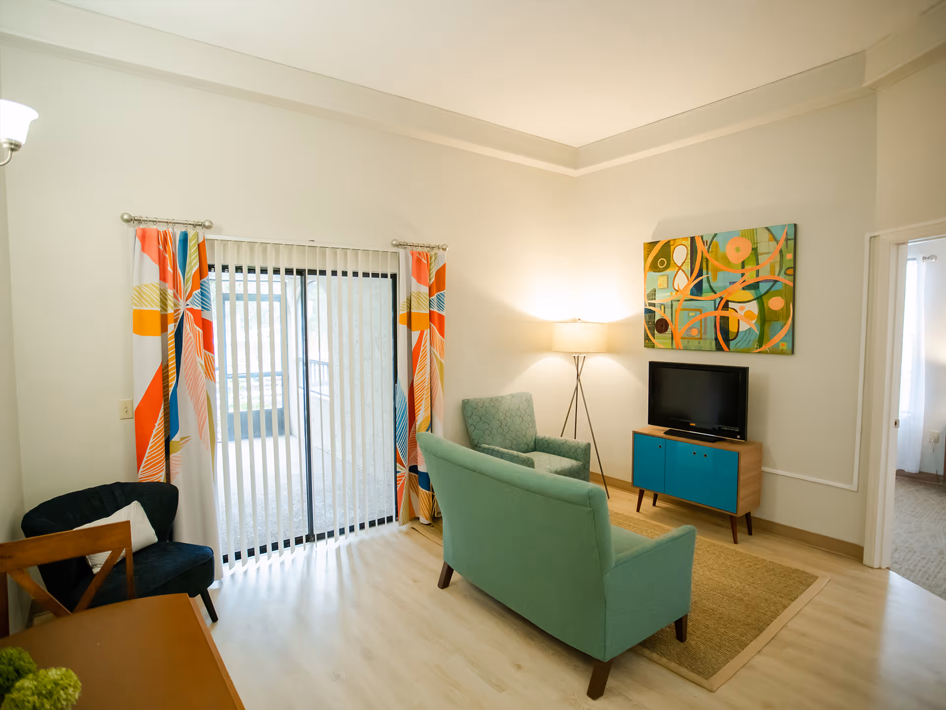 A cozy living room with light wood flooring, a teal loveseat and matching armchair, a small wooden TV stand with a flat-screen TV, a colorful abstract painting on the wall, a floor lamp, and sliding glass doors with vertical blinds and vibrant patterned curtains.