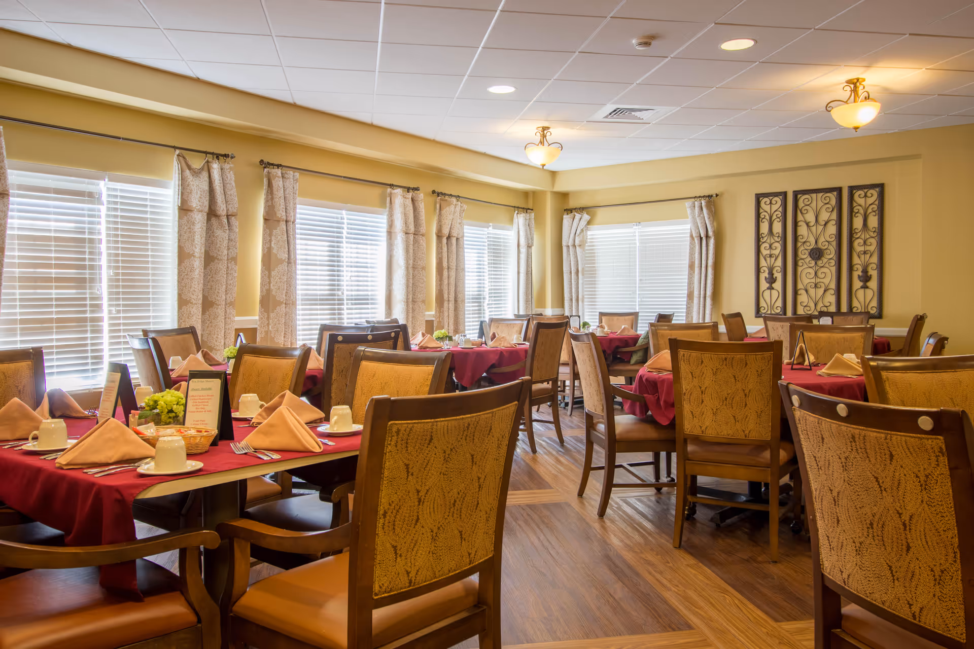 Dining room with multiple tables set with red tablecloths, folded napkins, place settings and wooden chairs beneath large windows.