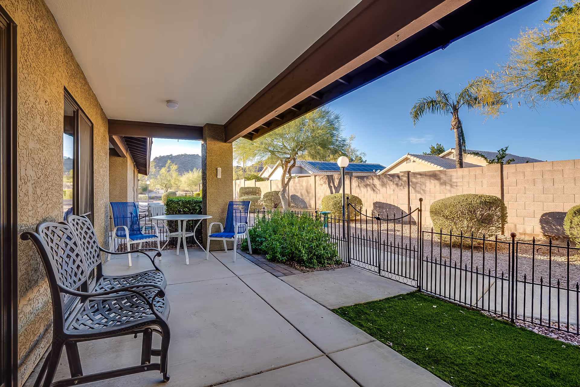 Covered patio area with a metal bench and a small round table surrounded by three blue chairs. The patio overlooks a fenced yard with green bushes, a palm tree, and a stone wall with houses and solar panels in the background under a clear blue sky.