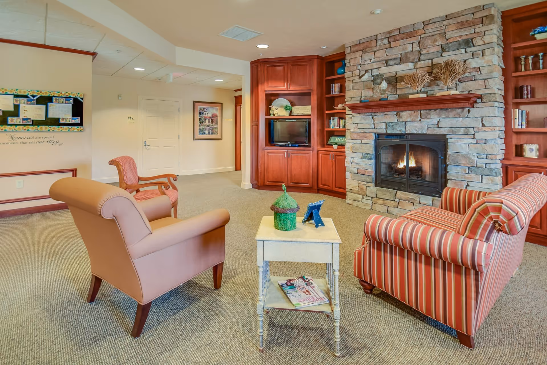 A cozy assisted living facility common area with a stone fireplace, built-in wooden shelves and cabinets, a small white side table with decorative items, and three upholstered chairs arranged around the table. The room has beige carpeting and light-colored walls with framed artwork and a bulletin board.