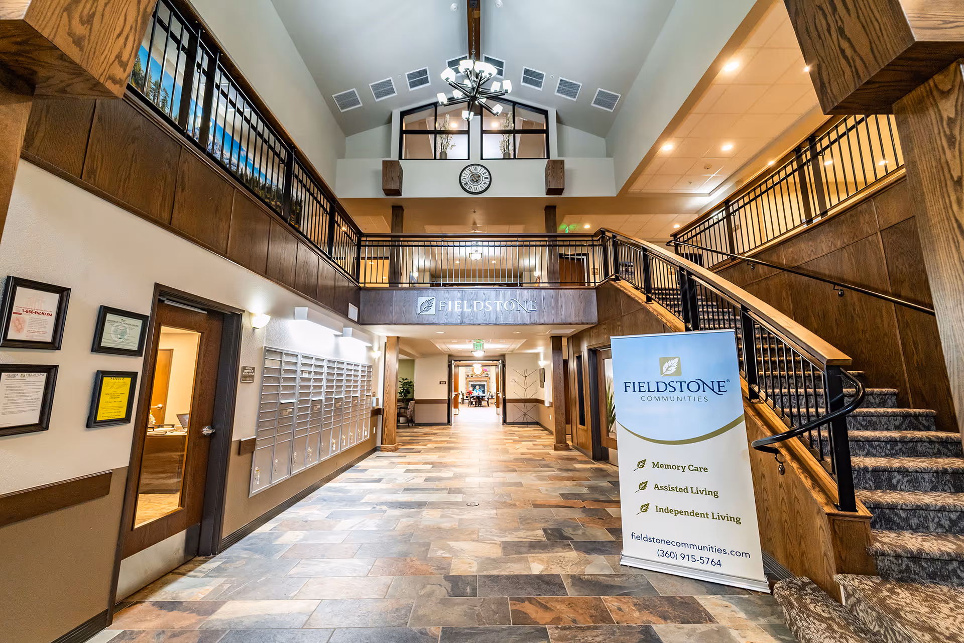 Interior view of Fieldstone Cooper Point senior living facility showing a spacious lobby with tiled floor, wooden staircase with black railings on the right, mailboxes on the left wall, a clock above the entrance to an upper level, and a standing banner displaying Fieldstone Communities services including Memory Care, Assisted Living, and Independent Living.
