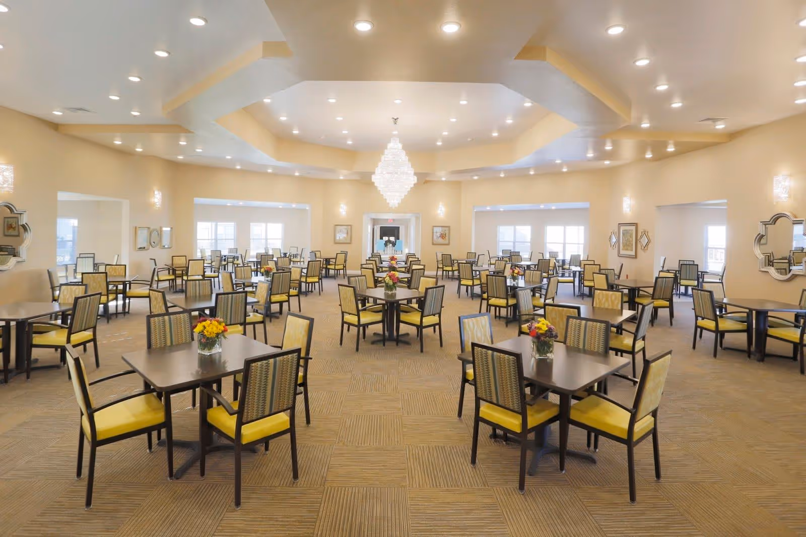 A spacious dining room with multiple square tables, each surrounded by four chairs with yellow cushions. The room features a high ceiling with recessed lighting and a large chandelier in the center. There are windows along the walls allowing natural light to enter, and decorative mirrors and framed artwork adorn the walls. Small flower arrangements are placed on each table.