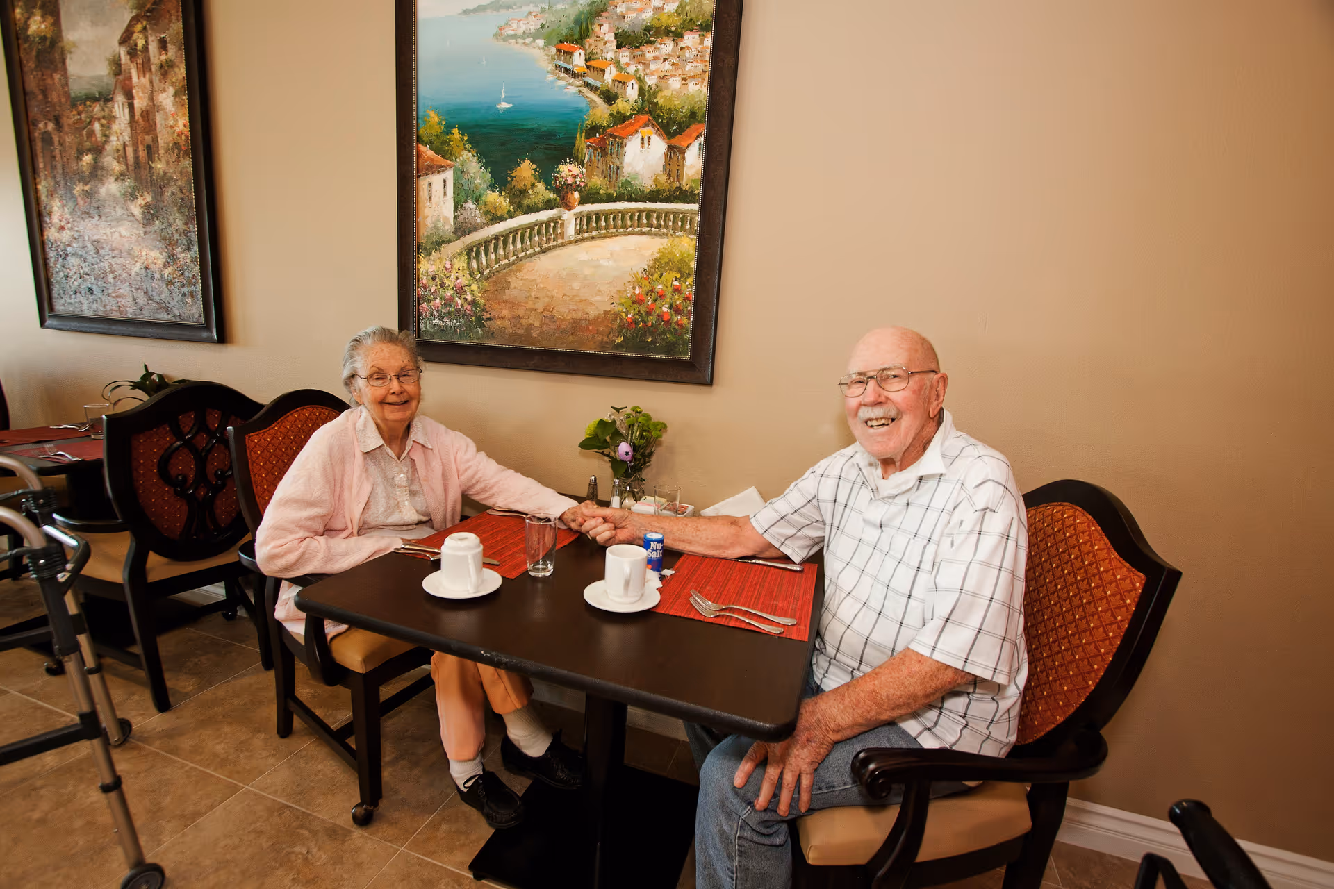An elderly couple sitting at a dining table in a senior living facility, holding hands and smiling. The table has two cups, glasses, and silverware on red placemats. Behind them are two framed paintings on a beige wall, and a walker is visible beside the woman.