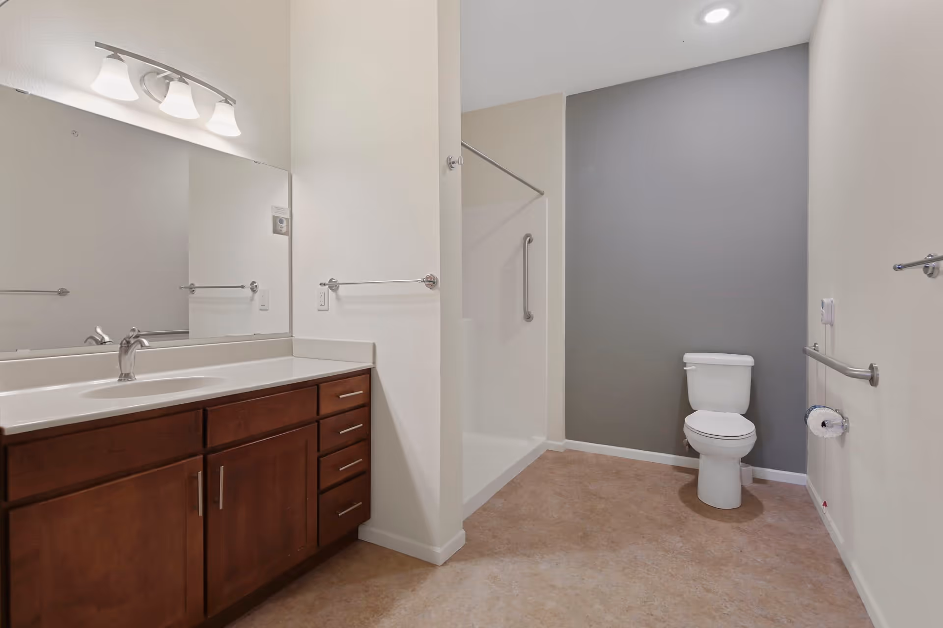 A spacious bathroom with a large mirror above a wooden vanity with a white countertop and sink. There is a walk-in shower with a grab bar and a toilet next to a gray accent wall. The floor is covered with beige tiles, and there are multiple grab bars on the walls for accessibility.