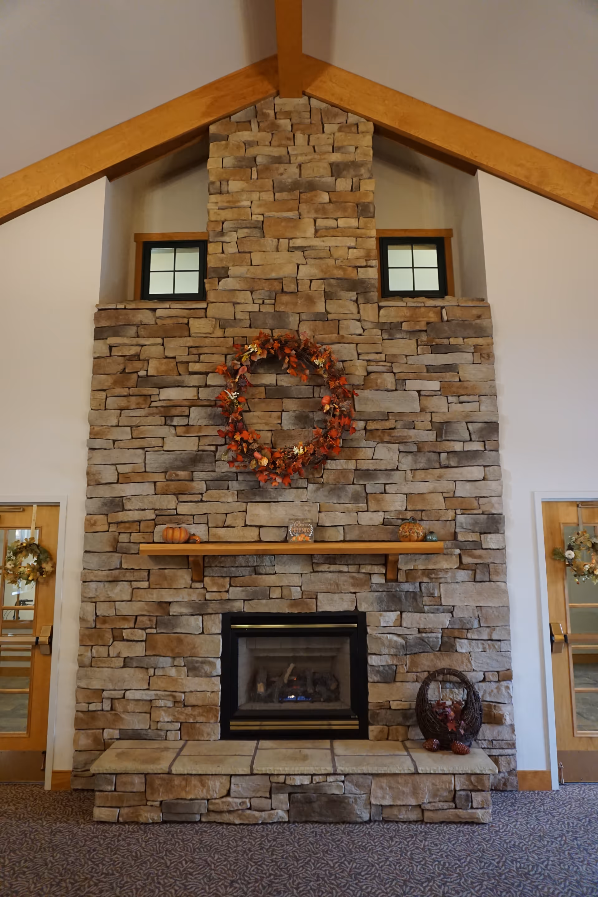 Large stone fireplace with a wooden mantel and autumn wreath in a vaulted-ceiling interior.
