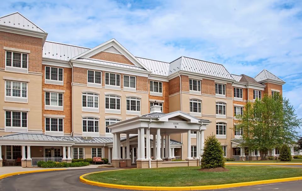 Front exterior of a four-story senior living facility with a covered porte-cochère, circular driveway, and landscaped lawn.