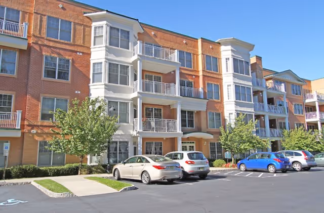 Brick three-story apartment building with white balconies and parked cars in front under a clear blue sky.