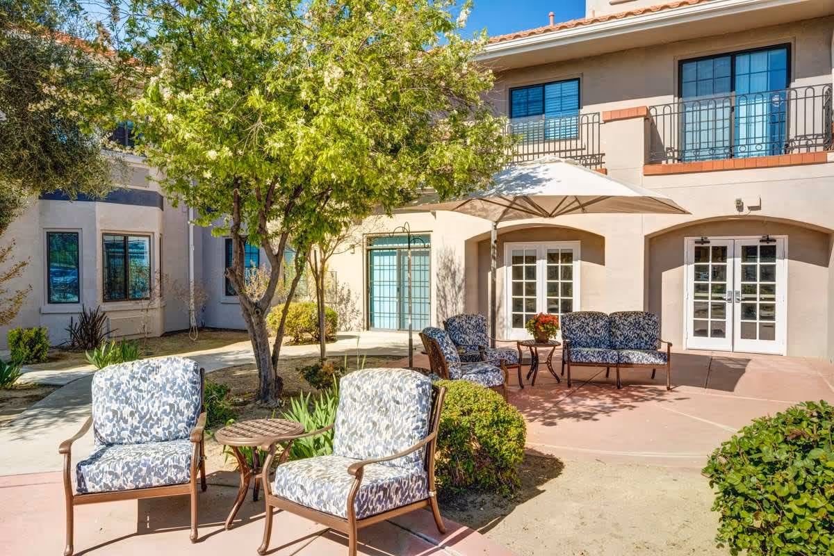Outdoor patio area at The Havens at Antelope Valley featuring cushioned chairs and a loveseat with a small table under a large umbrella, surrounded by trees and bushes, with a two-story building in the background.