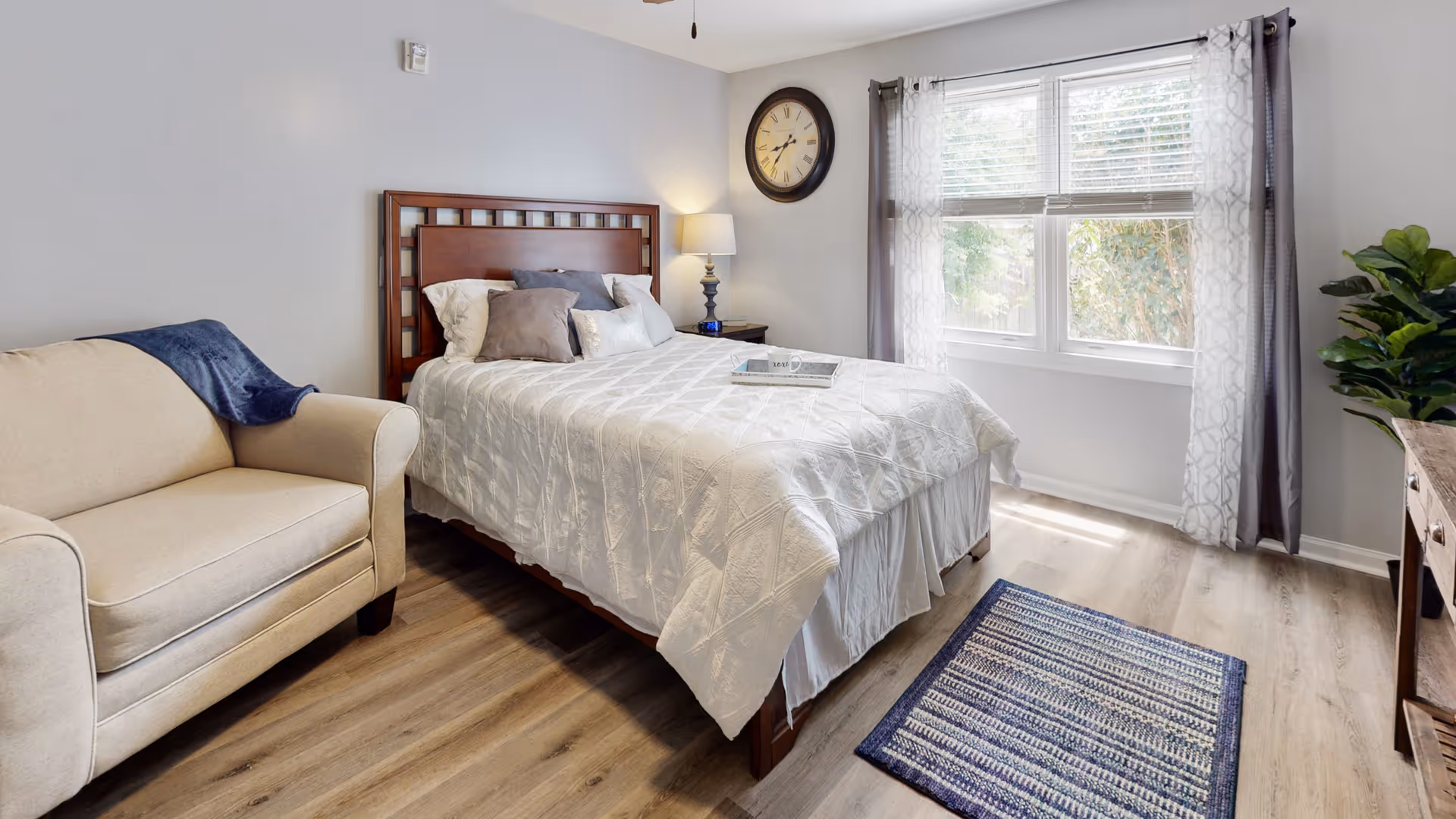A bright bedroom with a wooden bed frame, white quilted bedding, and multiple pillows. To the left of the bed is a beige armchair with a blue throw draped over the arm. A nightstand with a lamp and a large wall clock are on the far wall. A window with sheer and solid curtains lets in natural light. A blue and white patterned rug is on the wooden floor, and a green plant is on the right side near a wooden dresser.
