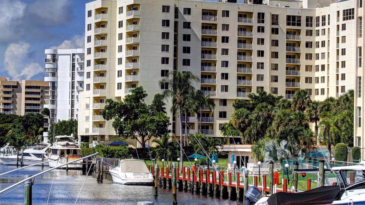 View of a waterfront residential building with multiple balconies, surrounded by palm trees and greenery. Several boats are docked along the water in front of the building under a partly cloudy sky.