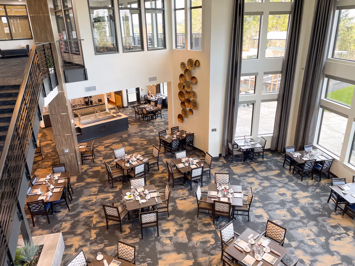 A spacious dining room with multiple tables set for meals, featuring chairs with patterned upholstery. The room has large floor-to-ceiling windows with gray curtains allowing natural light to fill the space. There is a decorative wall installation with circular metal pieces and a staircase with a metal railing on the left side. The dining area has a carpeted floor with a patterned design and a buffet station near the back wall.