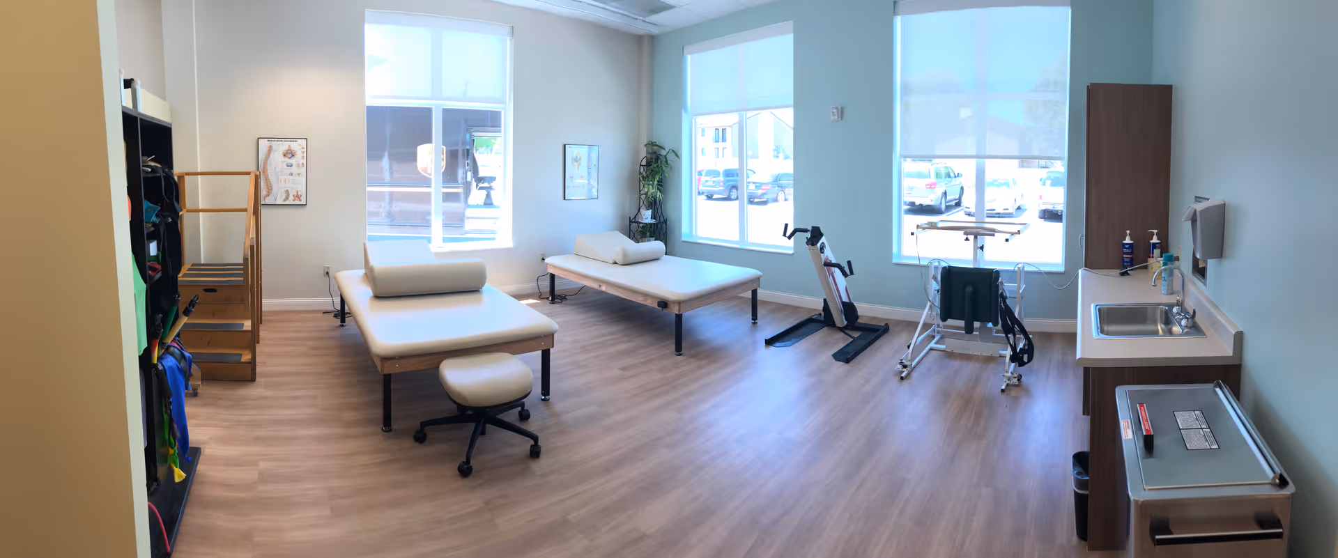 A bright rehabilitation therapy room with two padded treatment tables, a stool, exercise equipment including a stationary bike and parallel bars, a sink with hand sanitizer, and large windows letting in natural light.