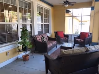 Covered outdoor patio area with wicker furniture including a sofa, two armchairs with red cushions, and a coffee table. The patio has a ceiling fan and is adjacent to a yellow exterior wall with multiple windows. There are potted plants on the floor and a small table.