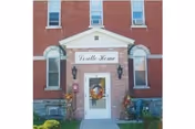 Front entrance of a red brick building with a white portico, a door with a wreath, and a sign above reading 'Loretto Home'.