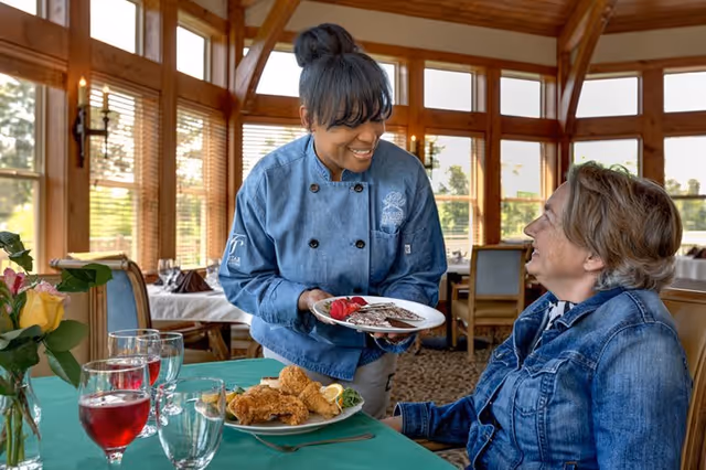 A smiling chef in a blue jacket serving a dessert plate to a seated woman in a denim jacket at a dining table with glasses of red beverage and a plate of fried chicken in a sunlit dining room with large windows and wooden beams.