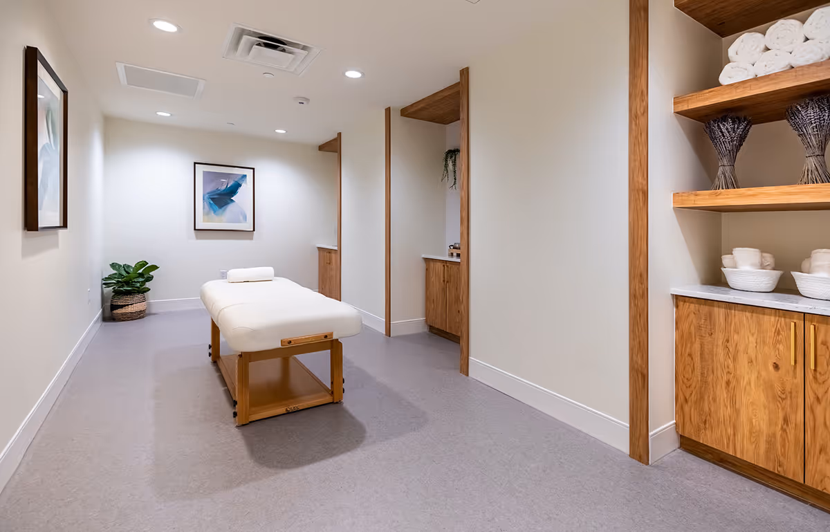 A clean and well-lit massage or therapy room with a padded massage table in the center, a rolled towel on the table, wooden shelves and cabinets holding rolled white towels and decorative lavender bundles, a potted plant in the corner, and framed abstract artwork on the walls.