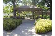 A shaded wooden gazebo surrounded by green bushes and trees with a concrete pathway leading to it, located in an outdoor garden area.