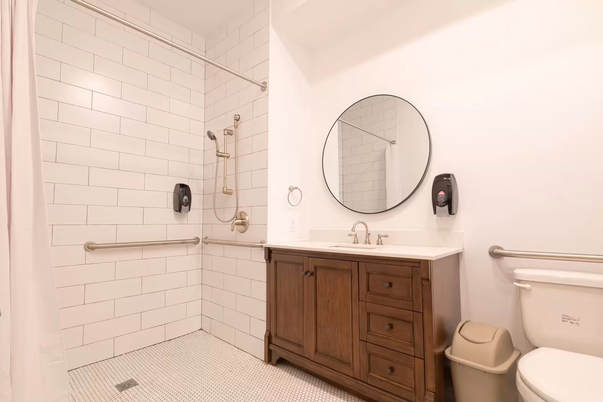 Bright accessible bathroom featuring a tiled walk-in shower with grab bars, a wooden vanity with round mirror, and a toilet.