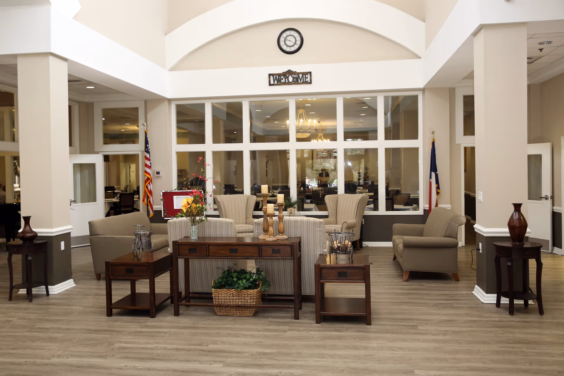 A spacious and well-lit living room area in a senior living facility with beige walls and wooden flooring. The room features comfortable armchairs and sofas arranged around wooden tables decorated with candles, flowers, and plants. Large windows and glass panels separate this area from another room in the background. An American flag and another flag are placed on either side of the room. A clock and a 'Welcome' sign are mounted on the wall above the windows.