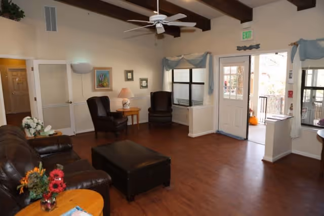 A cozy living room area in an assisted living facility with wooden flooring, a brown leather sofa, two dark armchairs, a black ottoman, a small wooden side table with a lamp, and a ceiling fan. The room has windows with light blue curtains and a door leading outside. There are framed pictures on the walls and a floor lamp in the corner.