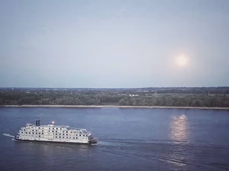 A white paddlewheel riverboat sails on a wide river at dusk with the moon reflecting on the water and a tree-lined shore in the distance.