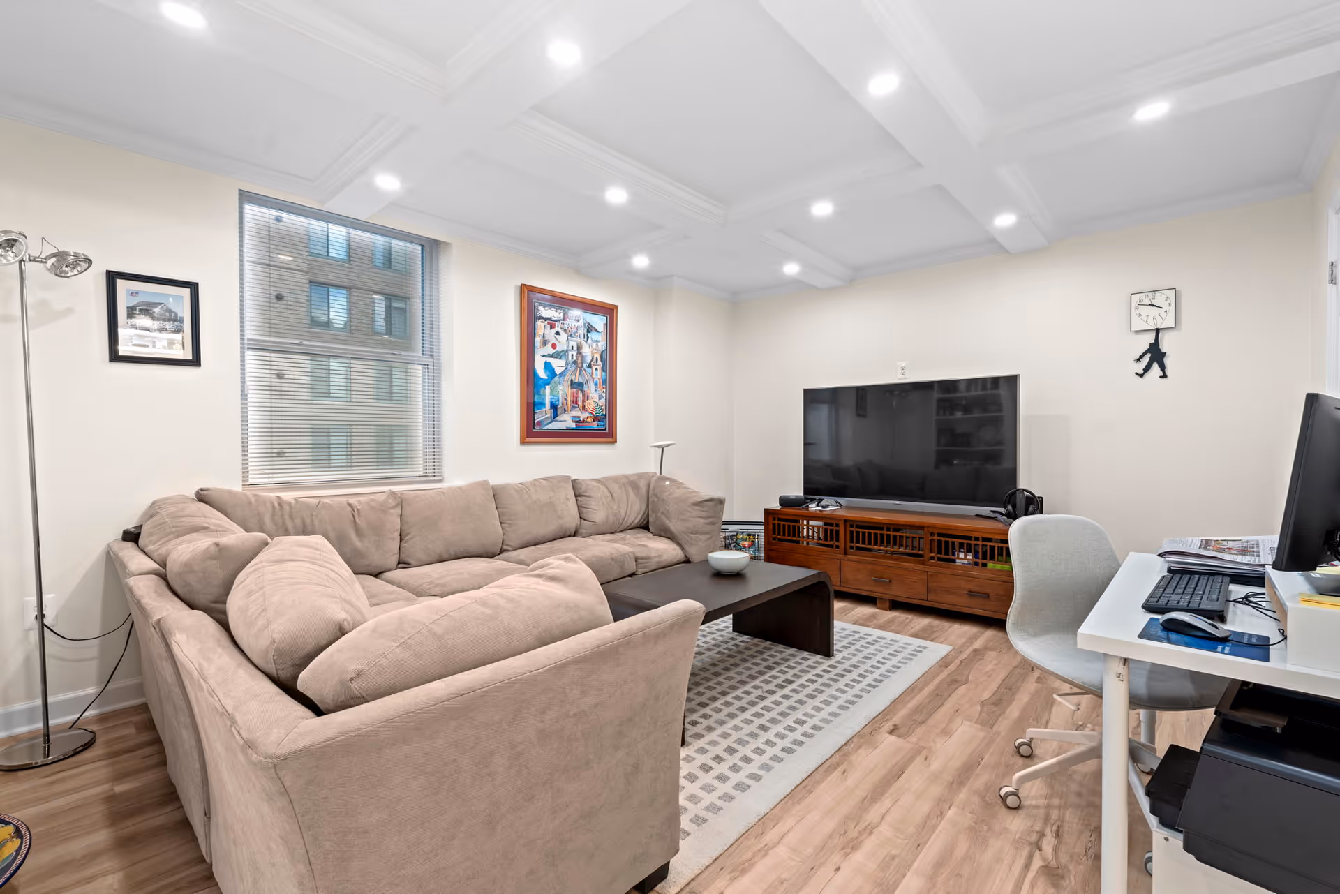 A cozy living room with a large beige sectional sofa, a dark wooden coffee table on a patterned rug, a flat-screen TV on a wooden media console, a white desk with a computer and office chair, and framed artwork on the walls. The room has recessed lighting and a window with blinds showing an exterior building.