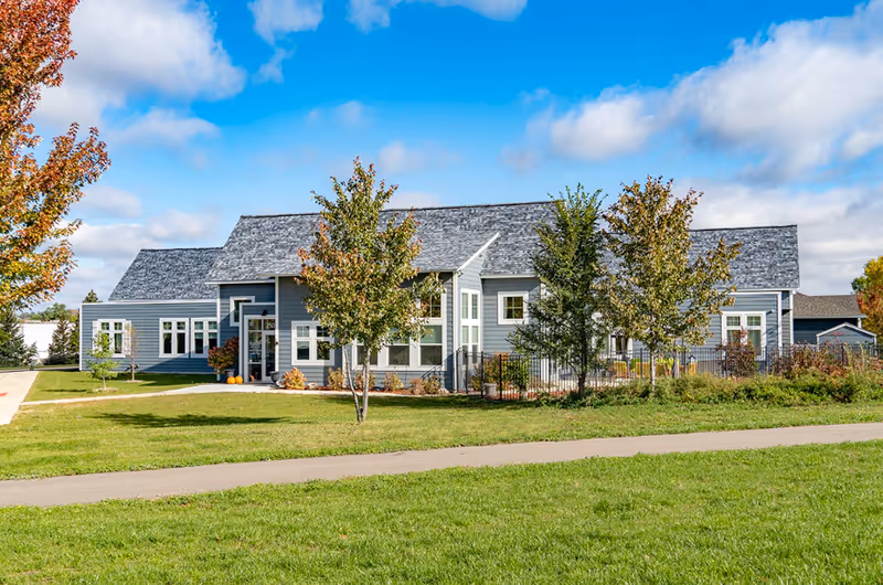 Single-story gray senior living building with a manicured lawn, trees, and a walkway under a blue sky.