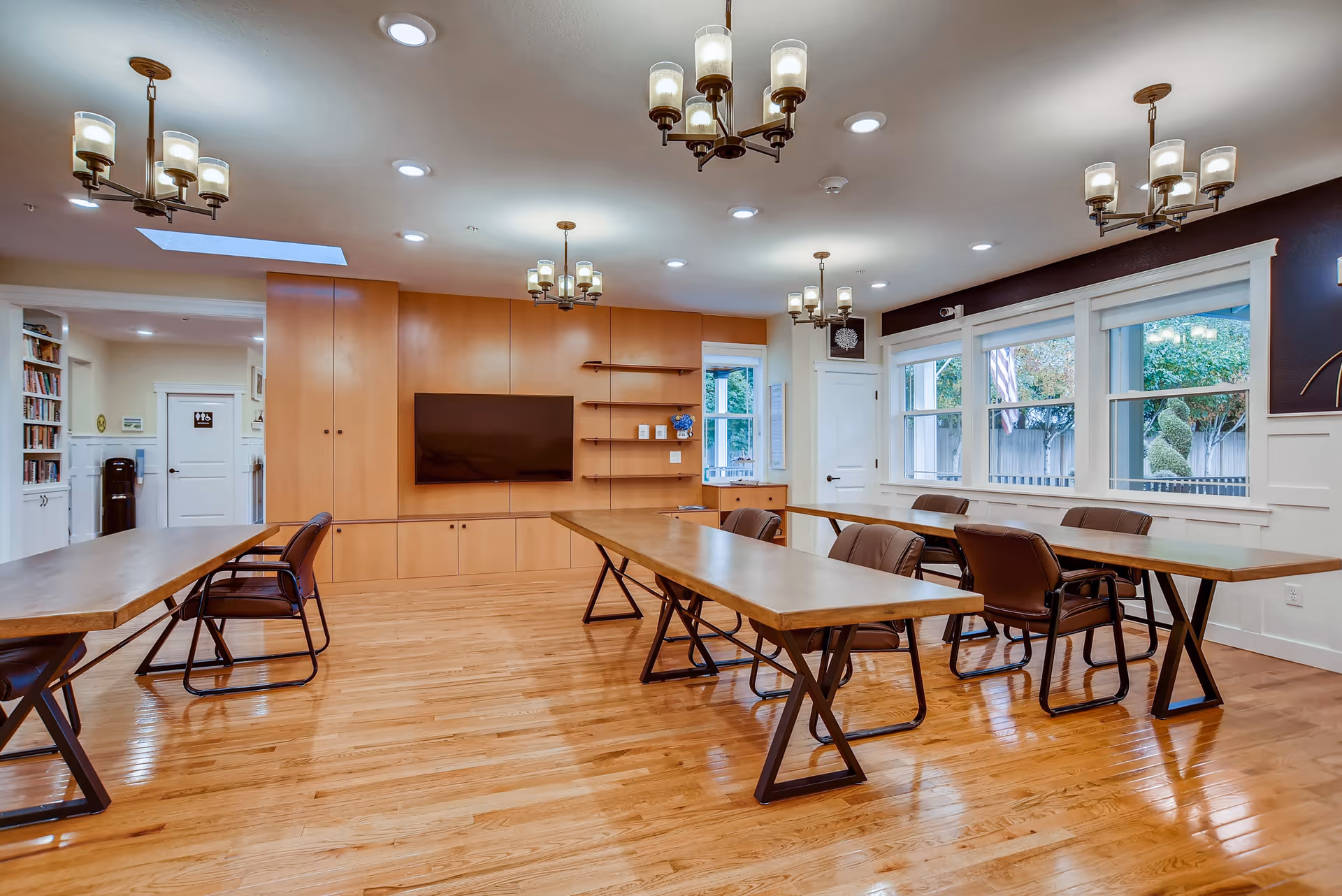 A spacious, well-lit common room with wooden floors and multiple rectangular wooden tables surrounded by brown chairs. The room features large windows with white trim letting in natural light, modern chandeliers hanging from the ceiling, and a wall-mounted flat-screen TV on a wooden panel wall with built-in shelves and cabinets. There is a water cooler and bookshelves visible in an adjacent room through an open doorway.