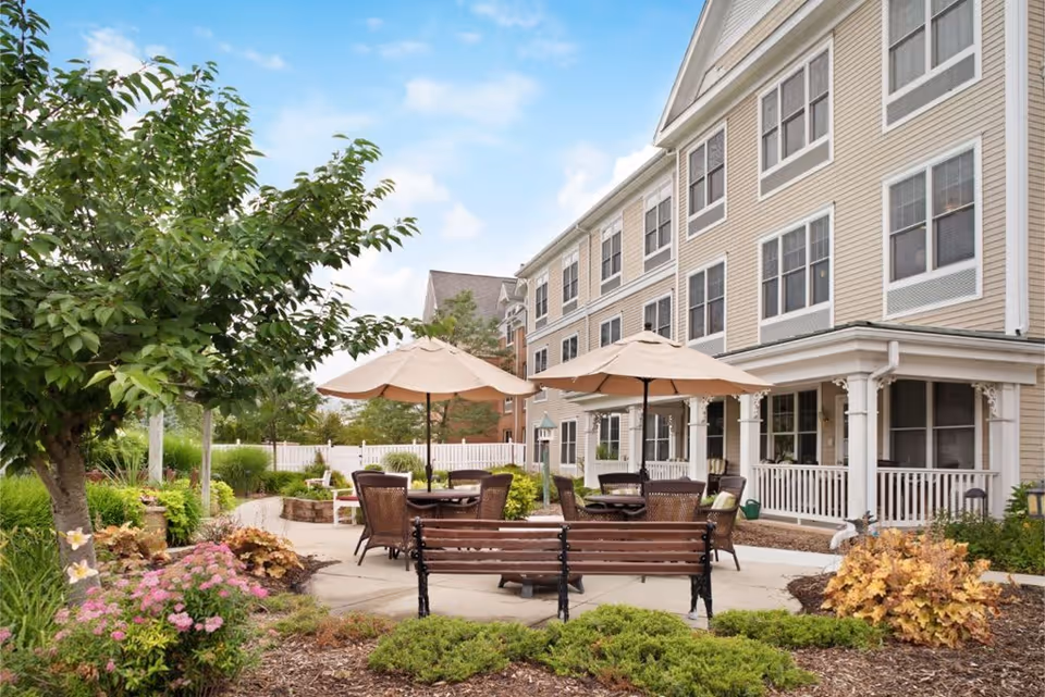 Outdoor patio area at a senior living facility with tables and chairs under large beige umbrellas, surrounded by landscaped greenery and flowers, with a multi-story beige building in the background under a partly cloudy sky.