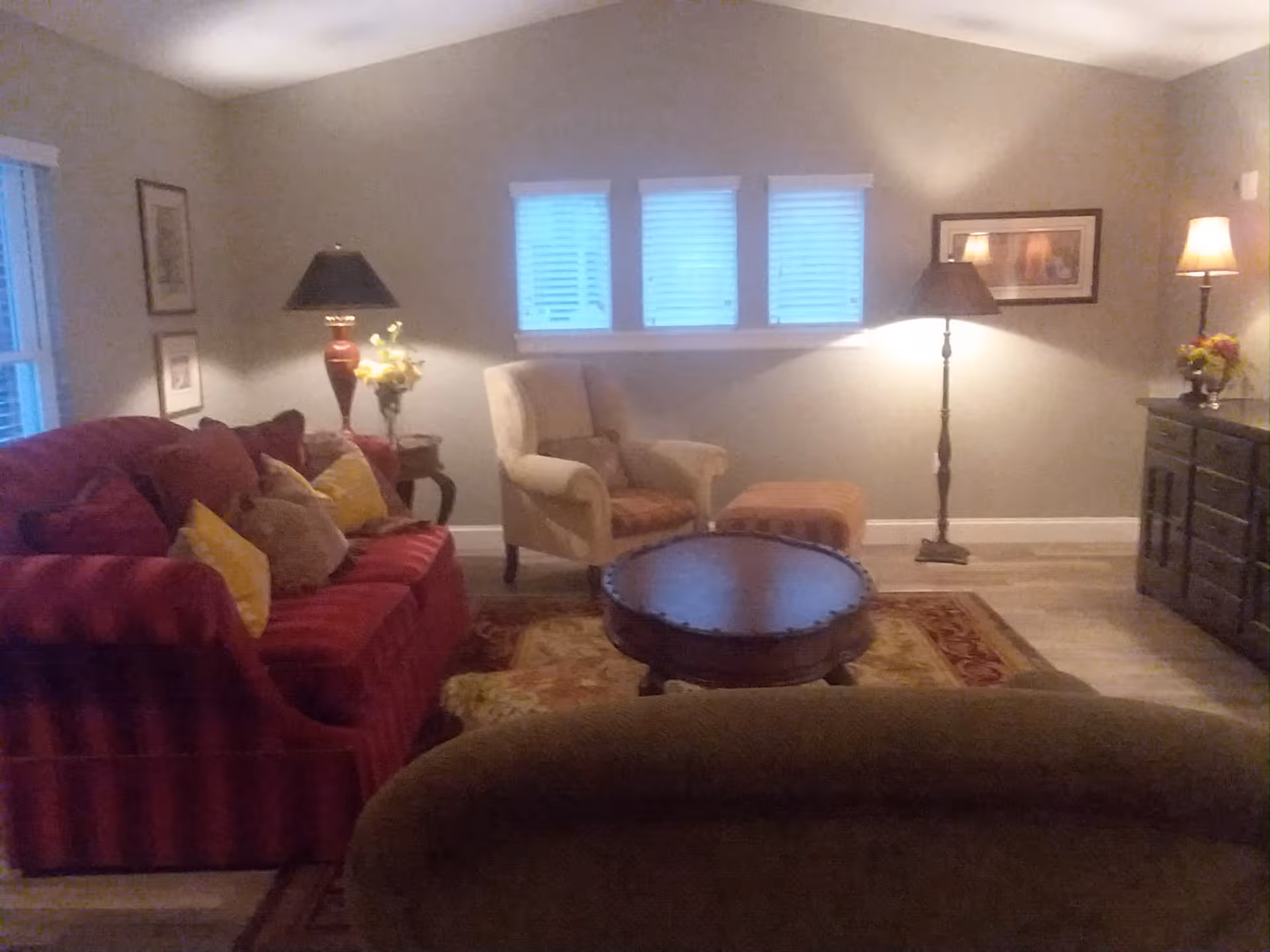 A cozy living room with a red sofa adorned with multiple cushions, a beige armchair with an ottoman, a round wooden coffee table, two floor lamps, and a side table with a flower vase. The walls are painted light gray and decorated with framed pictures. Three small windows with blinds are centered on the back wall.