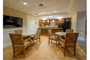 Communal dining area with several round tables and chairs in front of a kitchen/bar area and a wall-mounted TV.