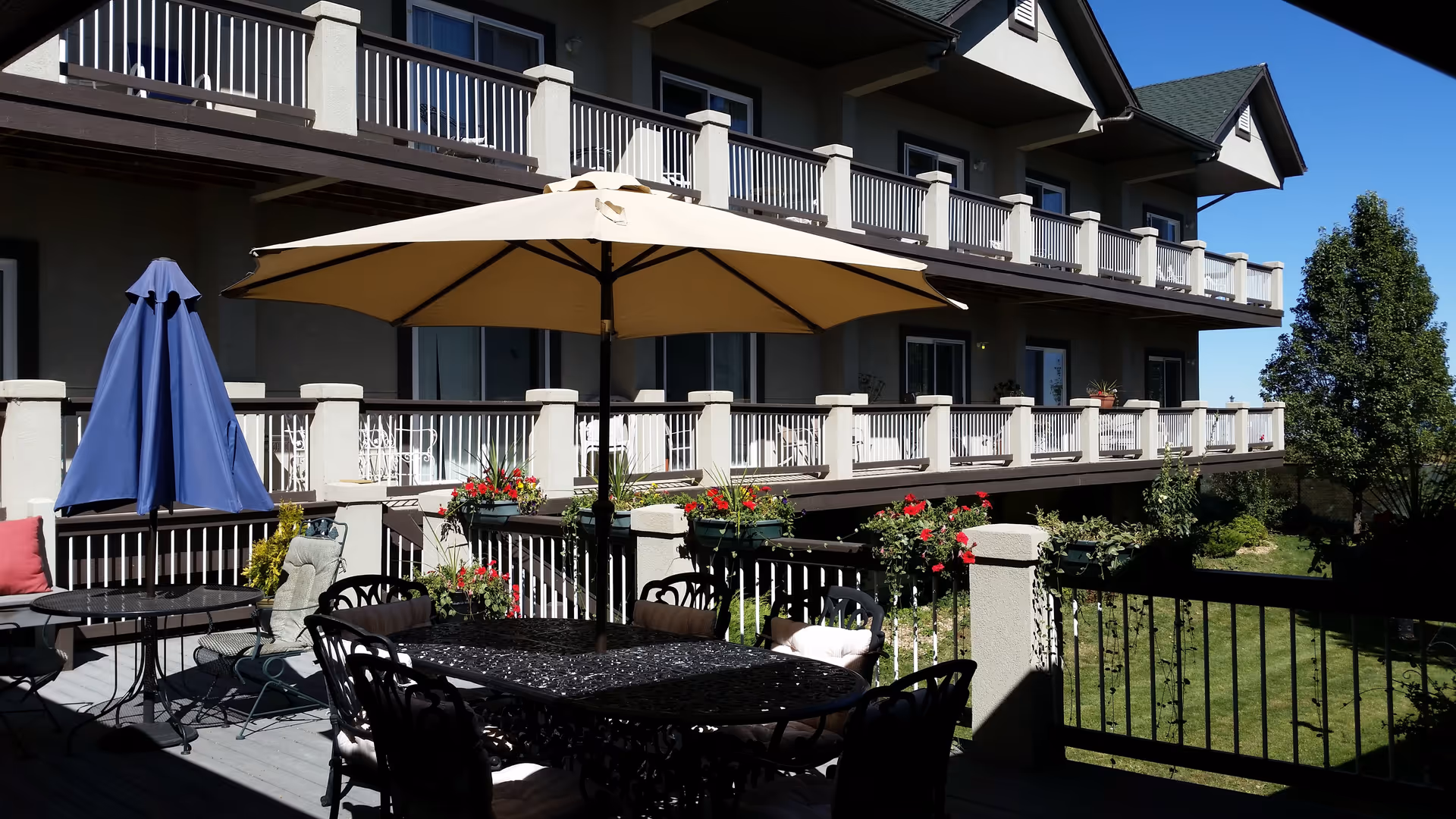 Outdoor patio area at The Manor on Marston Lake with a large beige umbrella over a black metal table surrounded by chairs. There is also a smaller table with a closed blue umbrella. The patio overlooks a green lawn with flower boxes containing red flowers attached to the railing. The building has two stories with balconies and white railings.