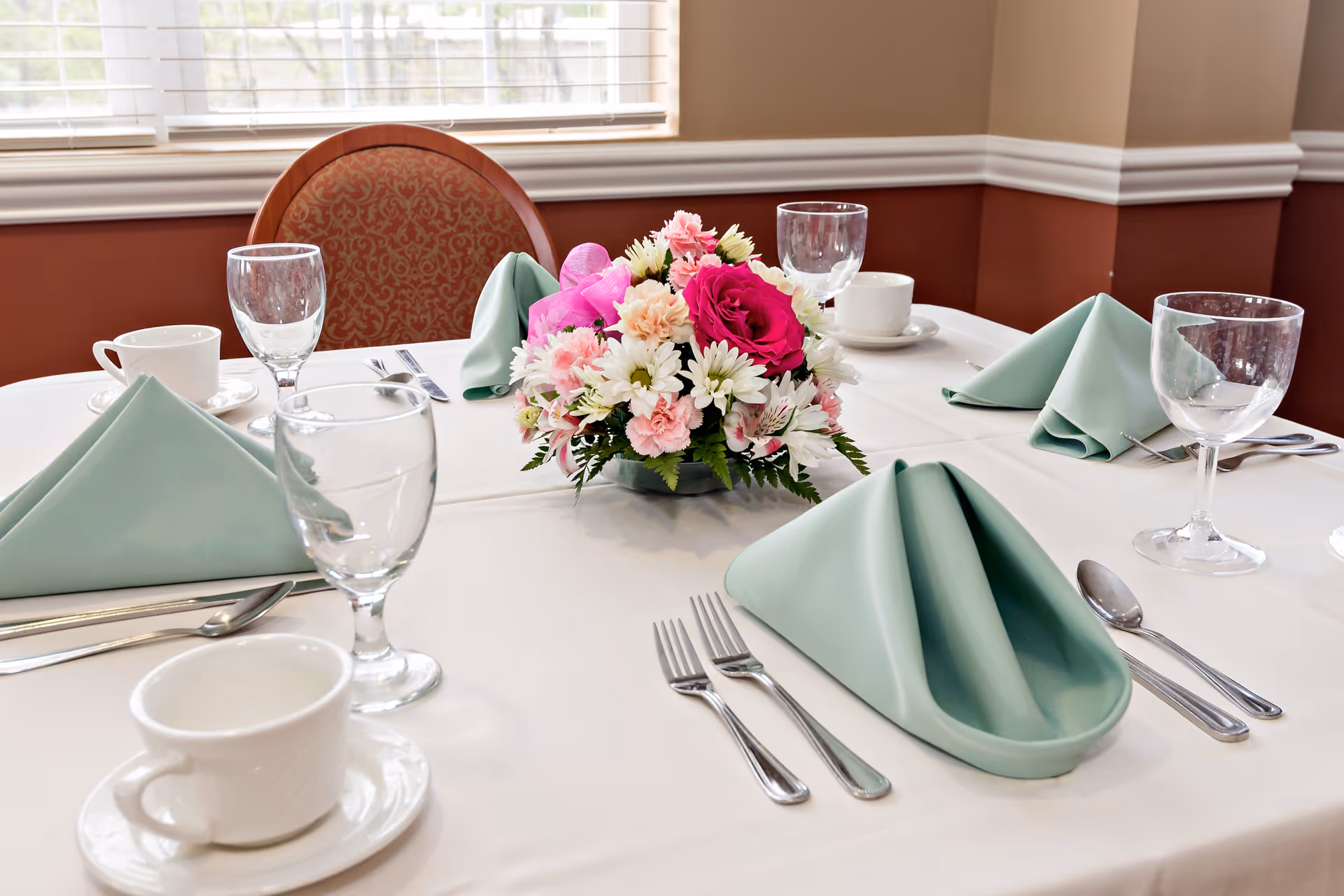 A dining table set with white tablecloth, light green folded napkins, clear water glasses, white coffee cups with saucers, silverware, and a centerpiece of pink, white, and red flowers. The background shows a window with blinds and a chair with a patterned backrest.