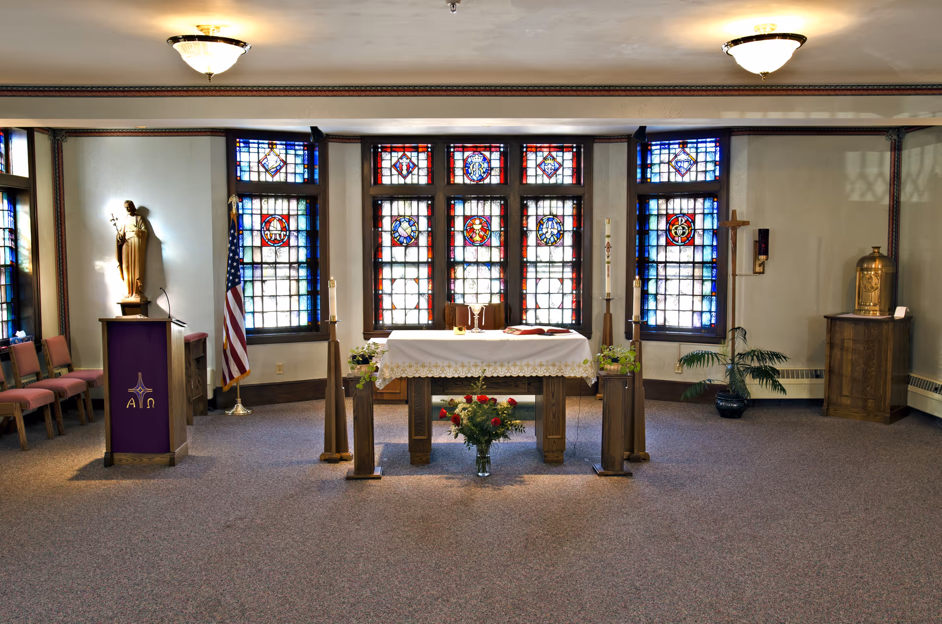 Interior view of a chapel with stained glass windows, an altar covered with a white cloth, a bouquet of red roses in front, a lectern with a purple cloth, a statue, an American flag, candles, and religious symbols.