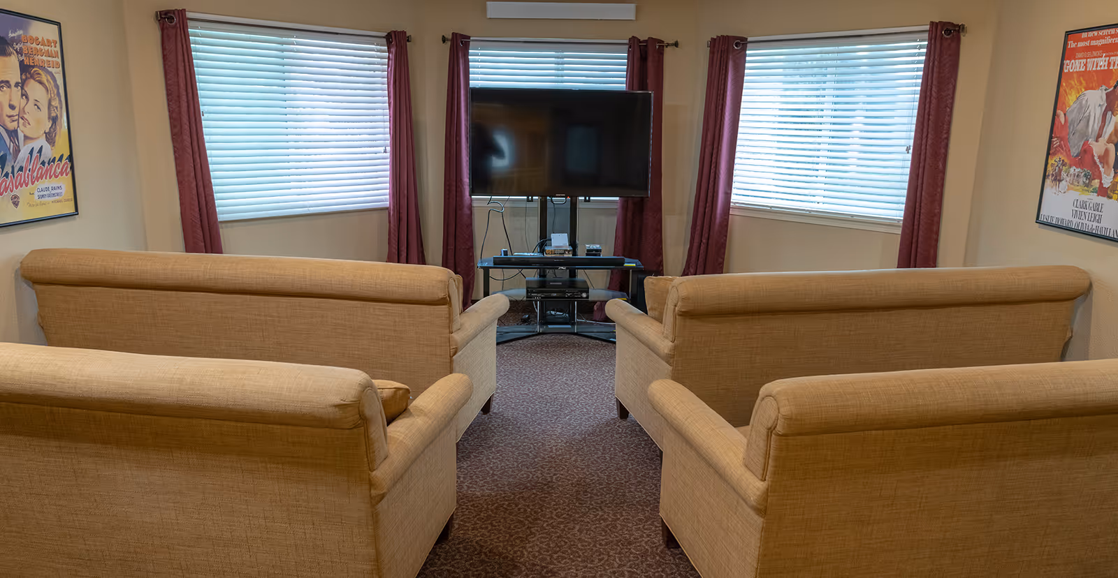 A small common living room with four beige sofas arranged facing a flat-screen TV set in front of three windows with blinds and red curtains.