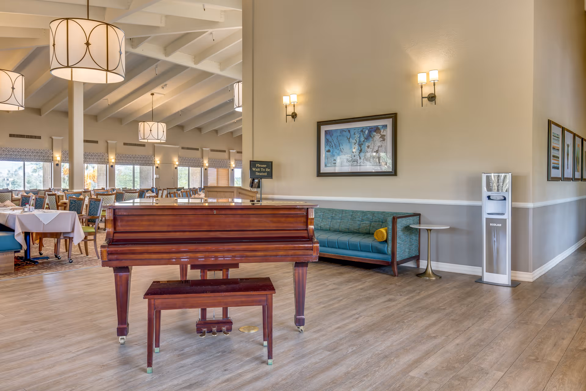 Interior view of a senior living facility dining area featuring a wooden piano with a matching bench in the foreground. The room has high vaulted ceilings with large hanging light fixtures, multiple dining tables with chairs, and a seating area with a green cushioned bench and a small round table. A water dispenser is positioned against the wall, which is decorated with framed artwork and wall sconces providing warm lighting.