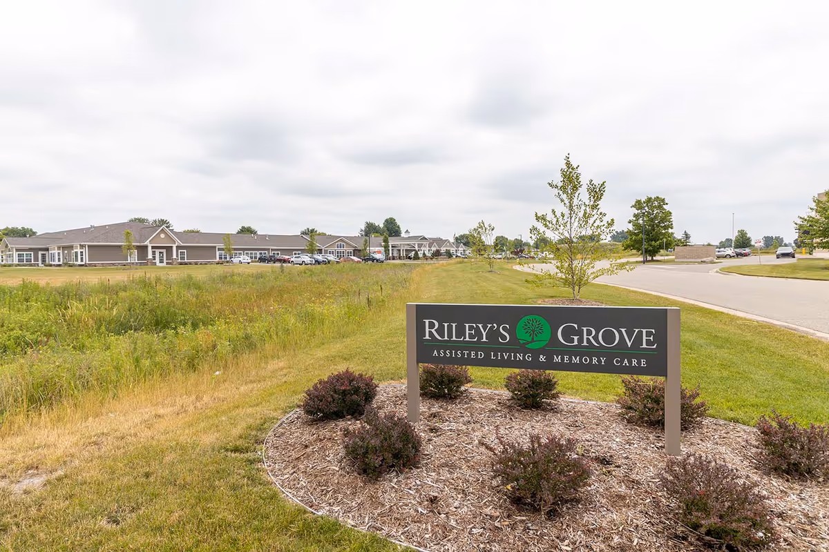 Exterior view of Riley's Grove Assisted Living & Memory Care facility with a large sign in the foreground surrounded by small bushes and a grassy area. The building and parking lot are visible in the background under a cloudy sky.