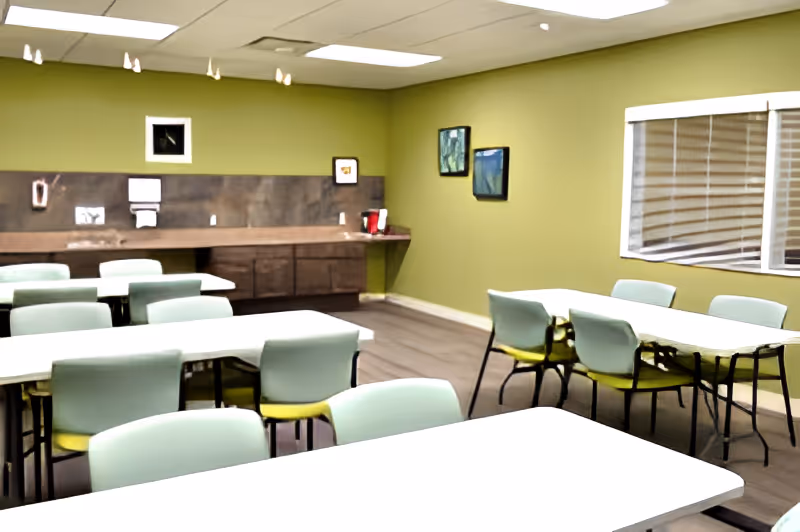 A room with multiple tables and chairs arranged for group seating, light green walls, a window with blinds, and a countertop with cabinets and a coffee maker in the back.