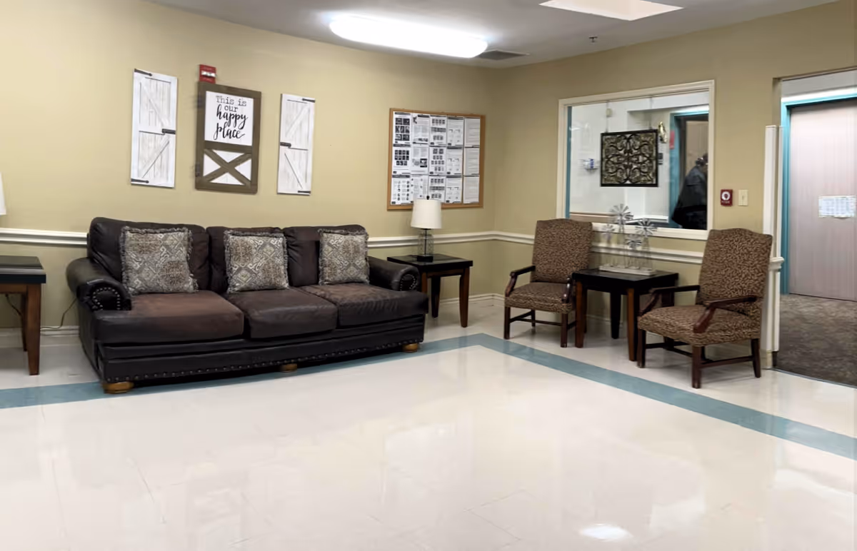 A waiting area in a nursing facility with a dark brown leather sofa adorned with three patterned cushions, two patterned armchairs, and two wooden side tables with lamps. The walls are beige with white trim, and there is a bulletin board and decorative wall art that says 'This is our happy place.' A window looks into another room, and a hallway with an elevator door is visible.