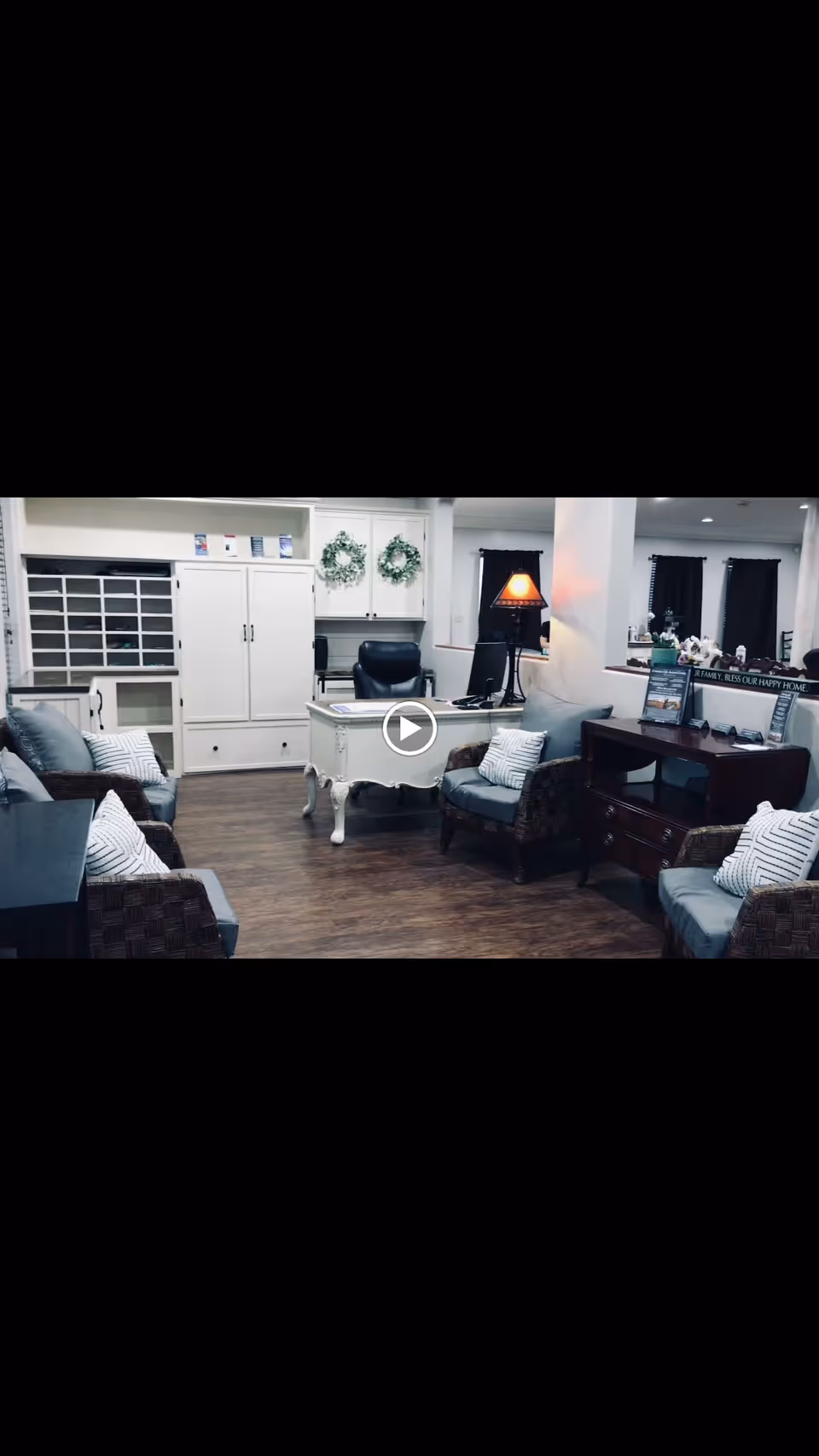 A cozy office or reception area with a white desk and black office chair in the center, surrounded by four cushioned armchairs with striped pillows. There is a wooden cabinet with drawers on the right side, a table lamp with a warm light, and decorative wreaths hanging on white cabinets in the background. The floor is wooden, and the space has a welcoming and comfortable atmosphere.