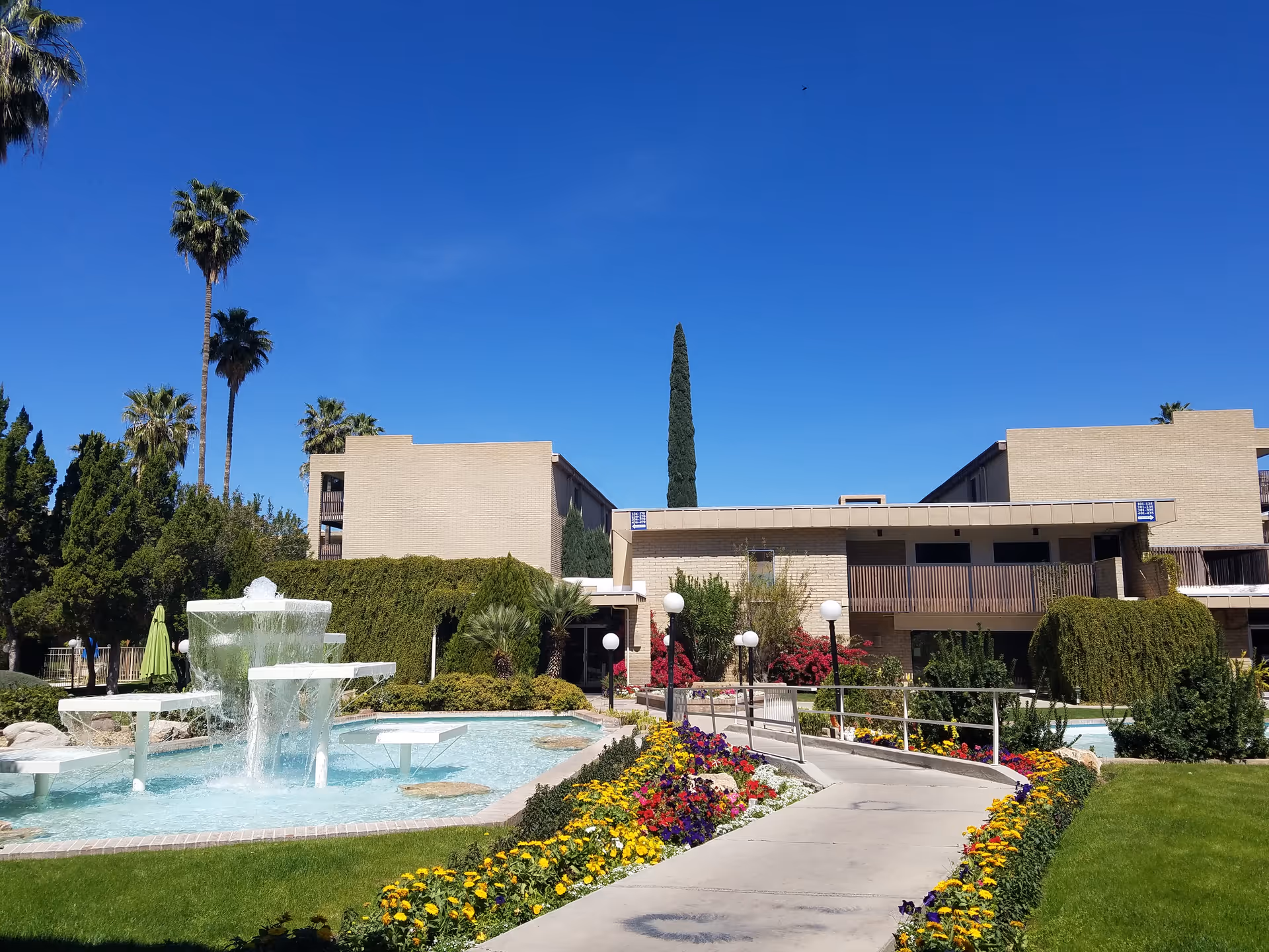 Outdoor view of Fellowship Square Tucson featuring a multi-tiered water fountain surrounded by a pool, a concrete walkway lined with colorful flowers, green grass, and various trees including tall palm trees. Beige buildings with balconies are visible in the background under a clear blue sky.