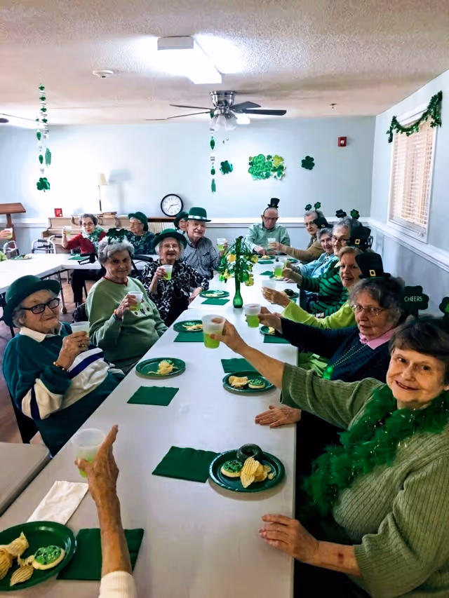 A group of elderly people sitting around a long table in a decorated room, raising green drinks in a toast. They are wearing green clothing and festive accessories, including hats and necklaces, celebrating a St. Patrick's Day themed event with green plates, napkins, and snacks on the table.