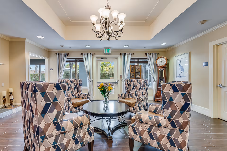 A cozy sitting area in a senior living facility with four patterned armchairs arranged around a round black coffee table holding a vase of flowers. The room has large windows with curtains, a grandfather clock, a framed bird artwork on the wall, and a chandelier hanging from the ceiling. The floor is tiled and there is an exit door in the background.