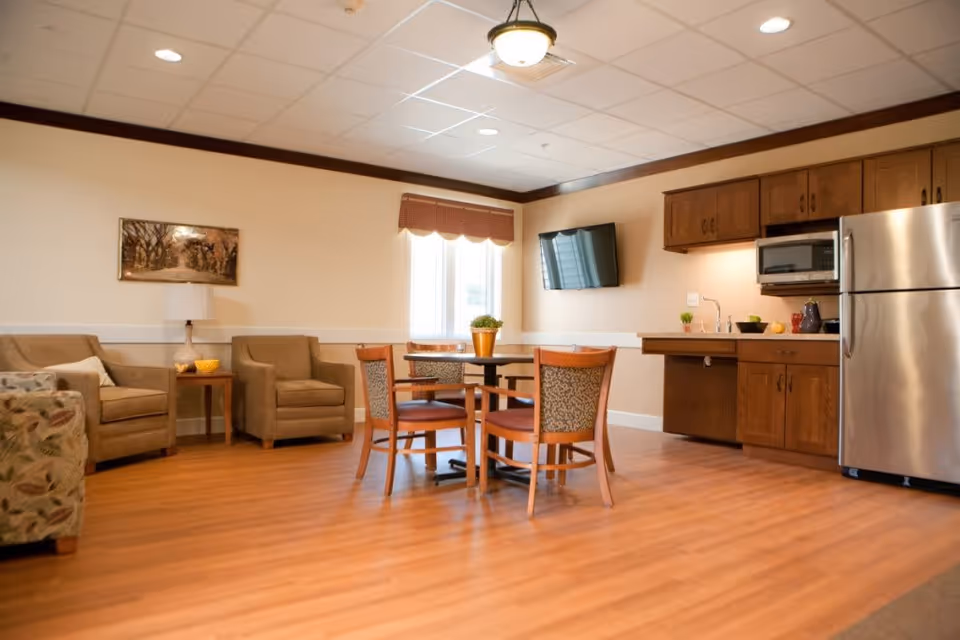 A cozy common area with a small kitchen and seating. The room features wooden flooring, a round table with four chairs, two armchairs with a side table and lamp, a wall-mounted TV, and kitchen cabinets with a microwave and stainless steel refrigerator. A window with a valance allows natural light into the space.