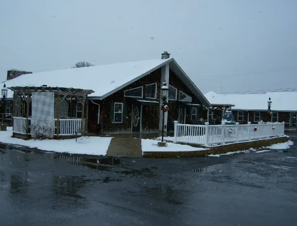 Exterior view of Fulmer's Personal Care Home building on a snowy day, showing a dark wooden structure with a peaked roof covered in snow, a white fence, and a wet driveway with some snow patches.