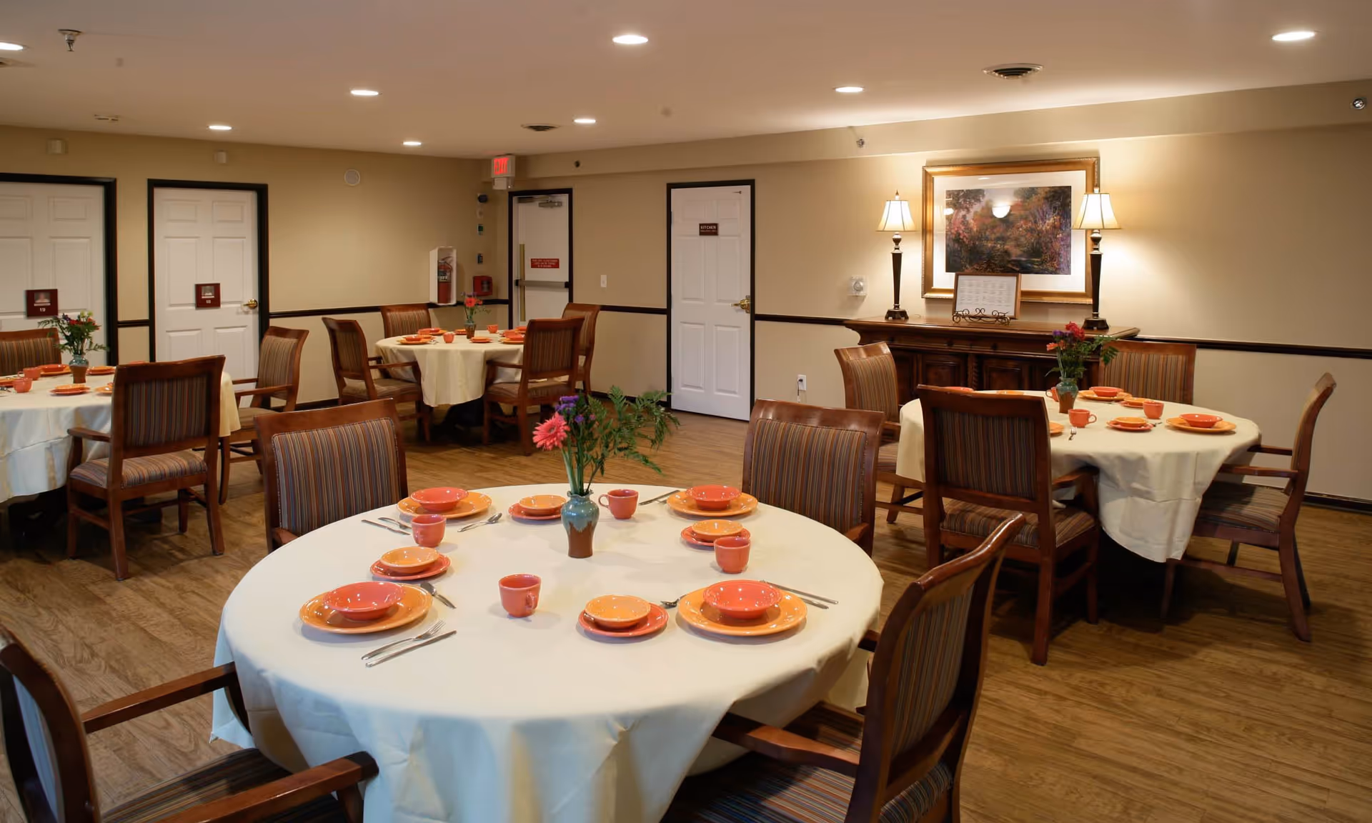 A dining room with round tables covered in white tablecloths, each set with orange plates, bowls, and cups. Wooden chairs with striped cushions surround the tables. The room has beige walls, wooden flooring, and a sideboard with two lamps and a framed painting above it.