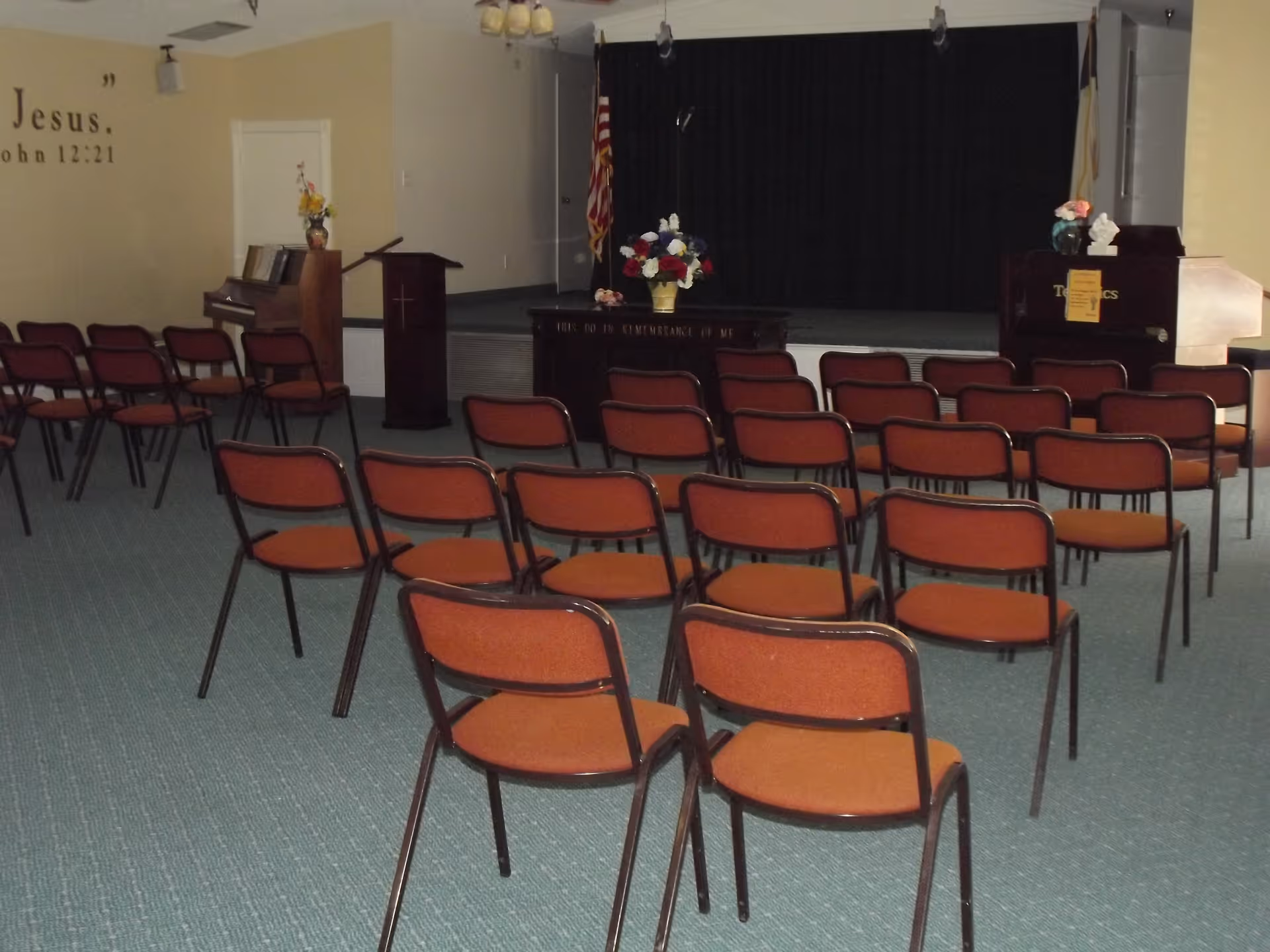 Rows of orange-upholstered chairs facing a small stage with podiums, a piano, flags, and floral arrangements in a chapel/meeting room.