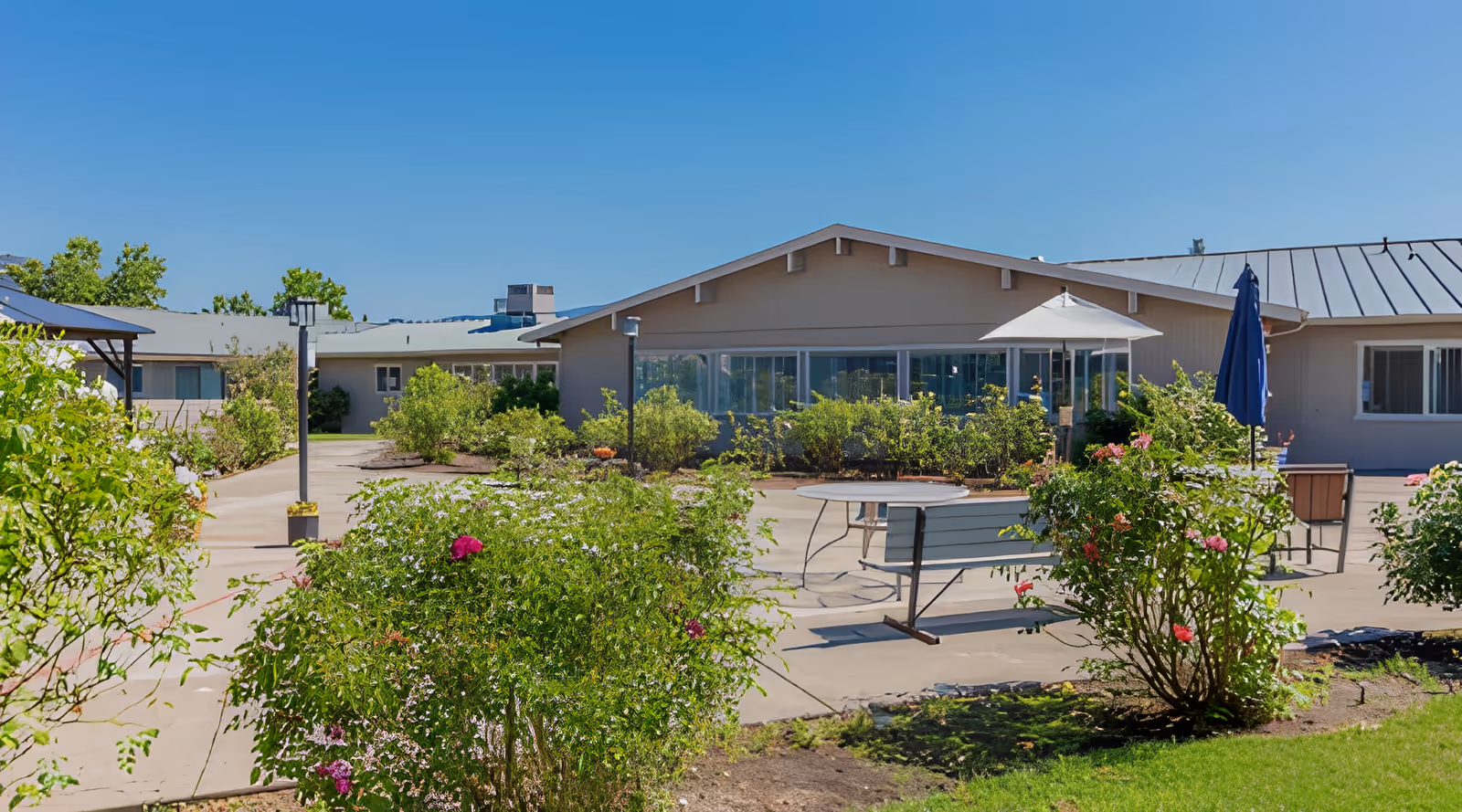 Sunny outdoor courtyard with benches, tables, umbrellas and landscaped shrubs in front of a single-story rehabilitation center building.
