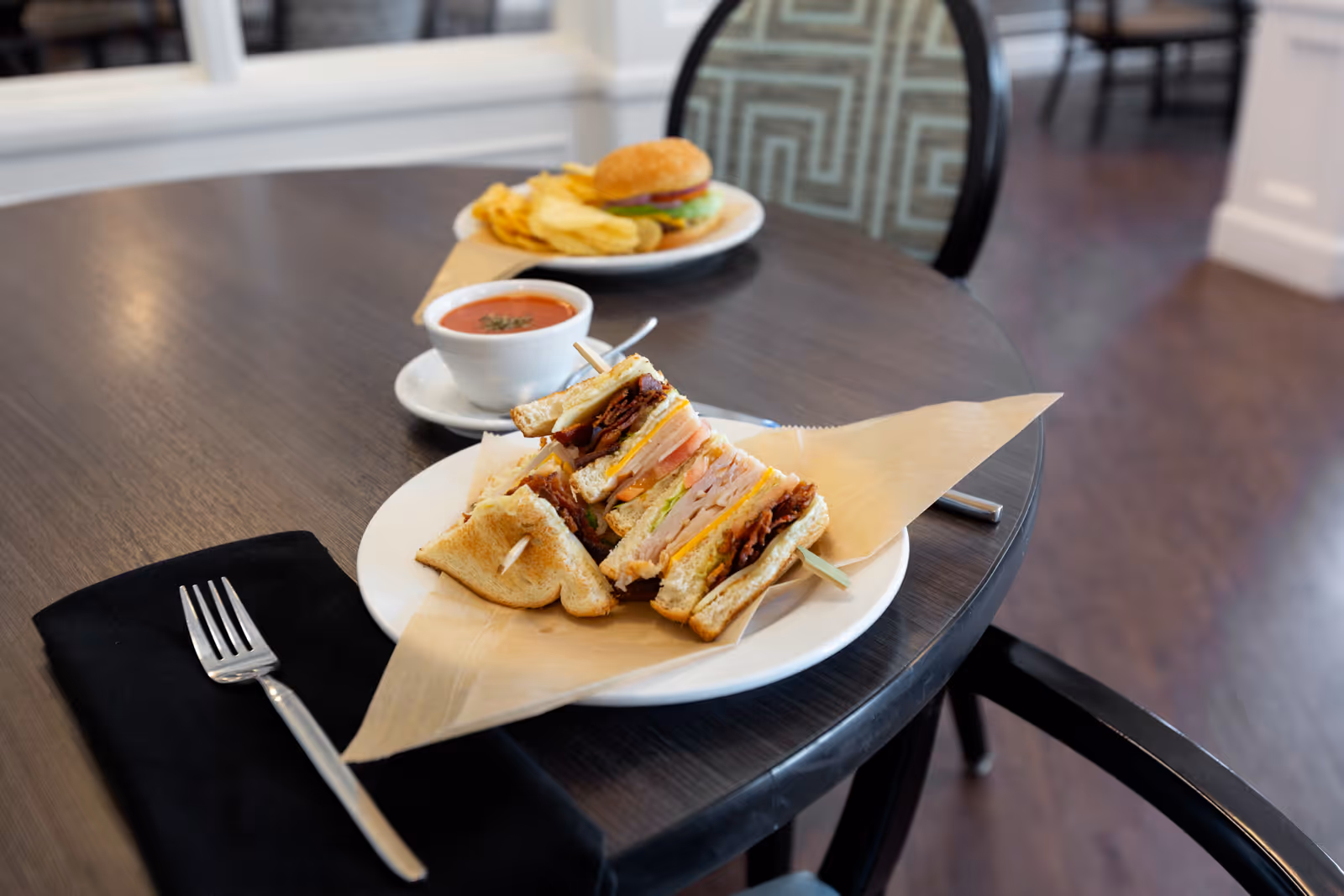 A close-up of a dining table with a club sandwich on a white plate, a cup of tomato soup, and a plate with a hamburger and potato chips in the background. A fork rests on a black napkin beside the sandwich. The setting appears to be a dining area with wooden flooring and patterned chairs.